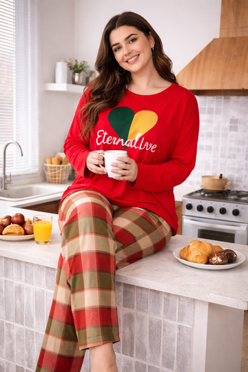 A woman sitting on a kitchen counter wearing a red long-sleeve shirt with a green and yellow heart graphic and the words 'Eternal Love' in white script, paired with red, green, and beige plaid pajama pants. She holds a white mug, with breakfast items on the counter in a bright kitchen setting.