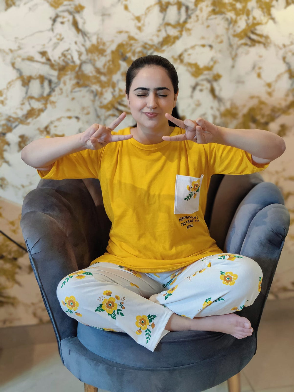 Person wearing a yellow shirt and floral pants sitting on a gray chair with a decorative wall in the background.